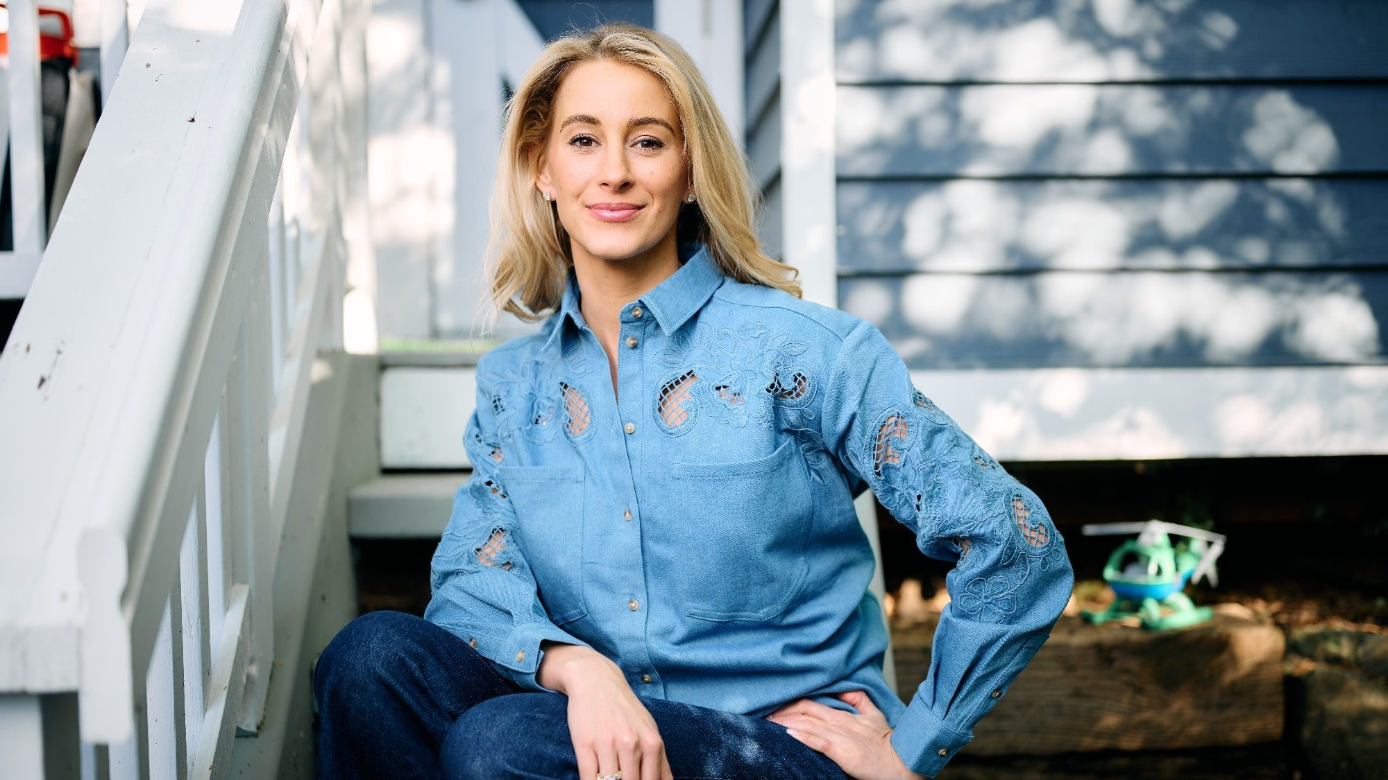 A woman in a blue denim shirt sits on wooden steps outside a house, left to her own devices, smiling at the camera.