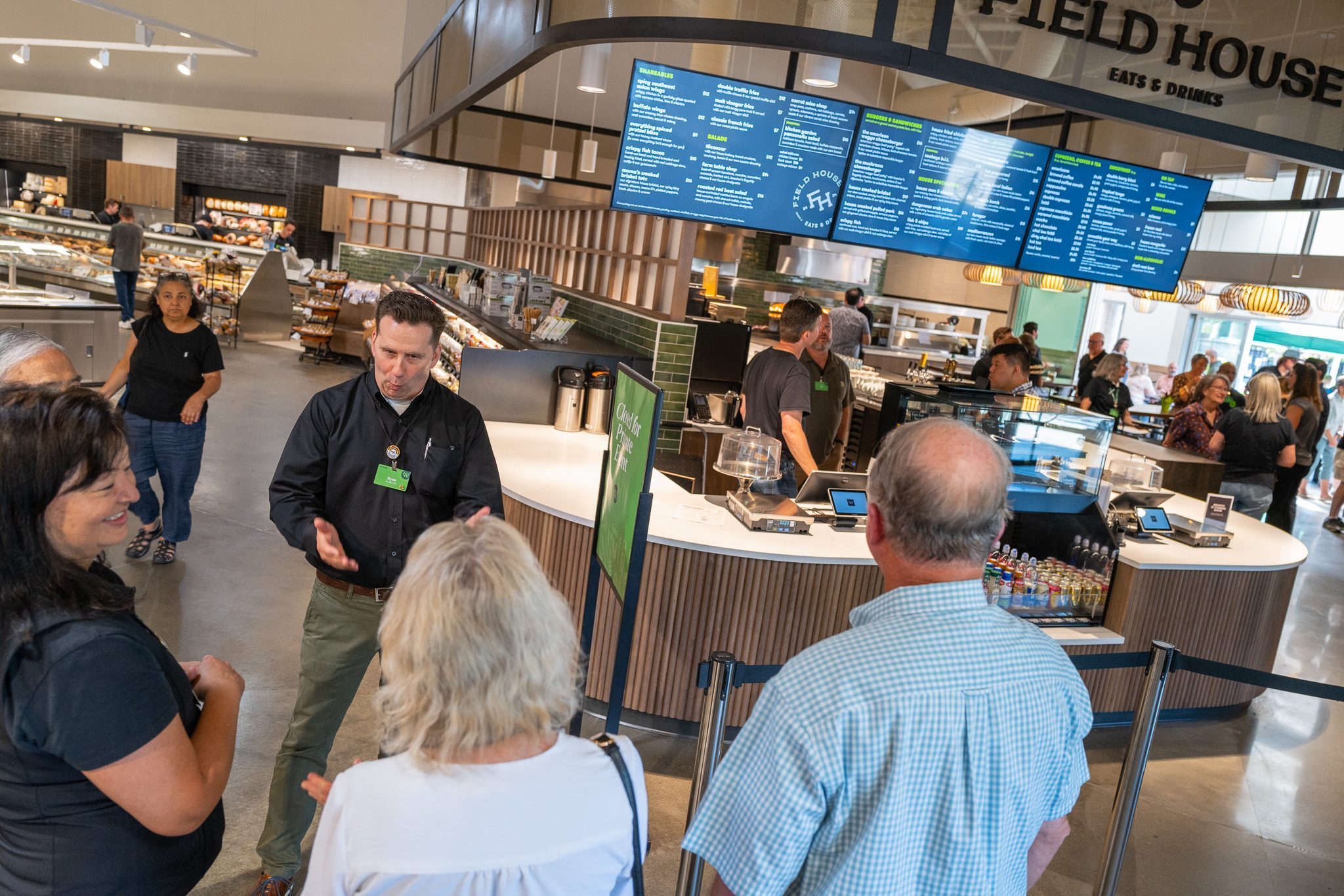 A group of people stand near a counter at "Field House Eats & Drinks" inside a food hall, engaging with staff members. A digital menu board and various food items are visible in the background.