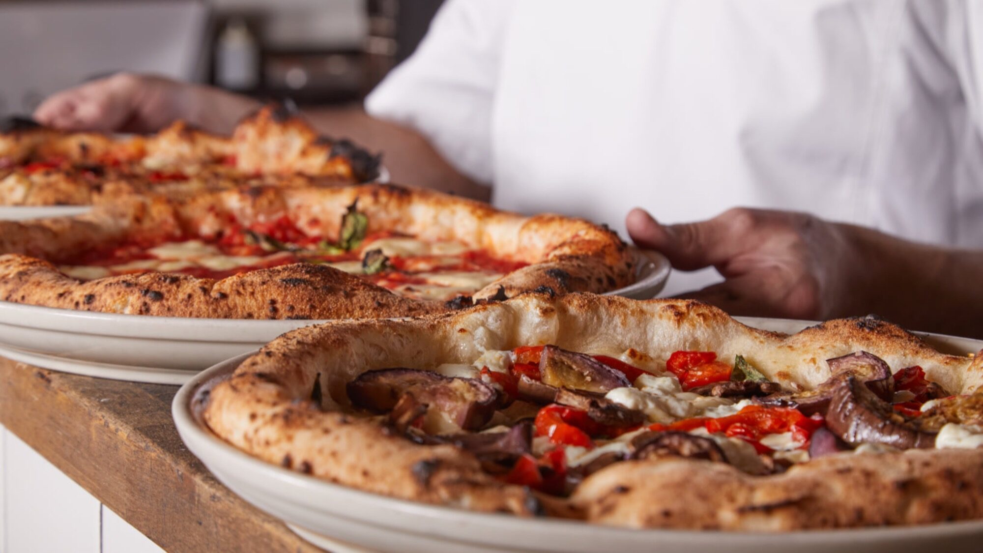 Two freshly baked pizzas with various toppings are served on white plates by a person in a white shirt.