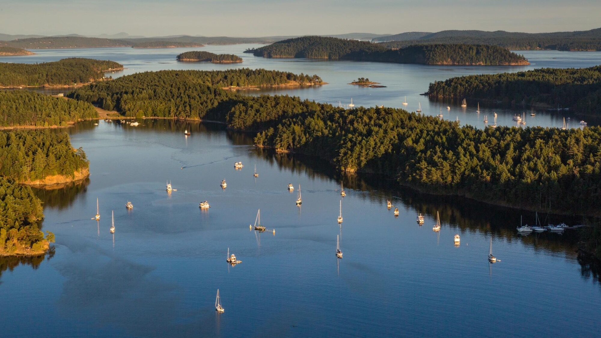 Aerial view of a coastal area with forested islands and numerous sailboats anchored in calm blue water under clear skies.