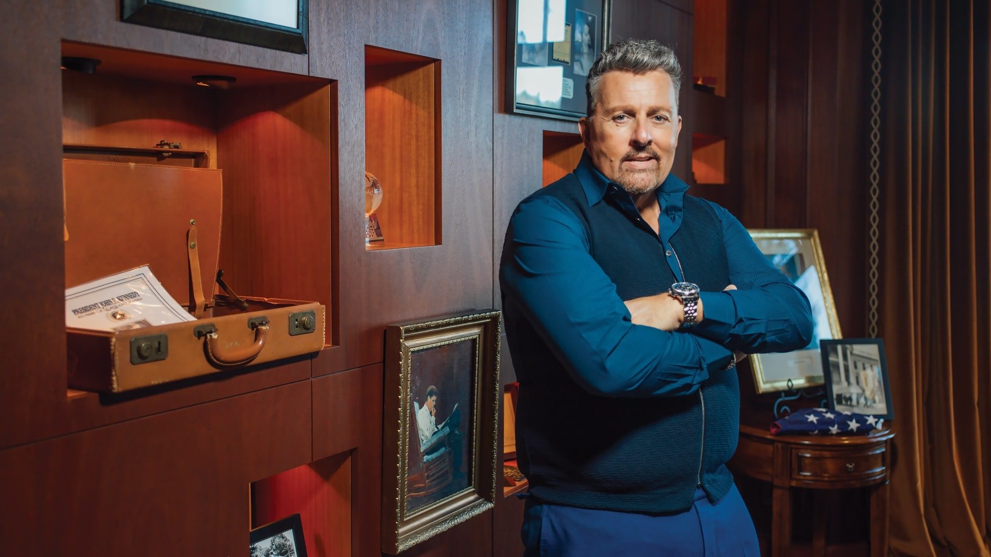 A man with folded arms stands in front of a wooden bookshelf displaying framed pictures, a leather suitcase, and various decorative items, evoking the charm of the Land Of Enchantment.