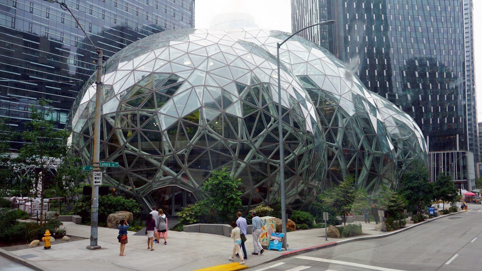 A geodesic dome structure with glass panels, surrounded by urban skyscrapers, with pedestrians walking on the sidewalk nearby.