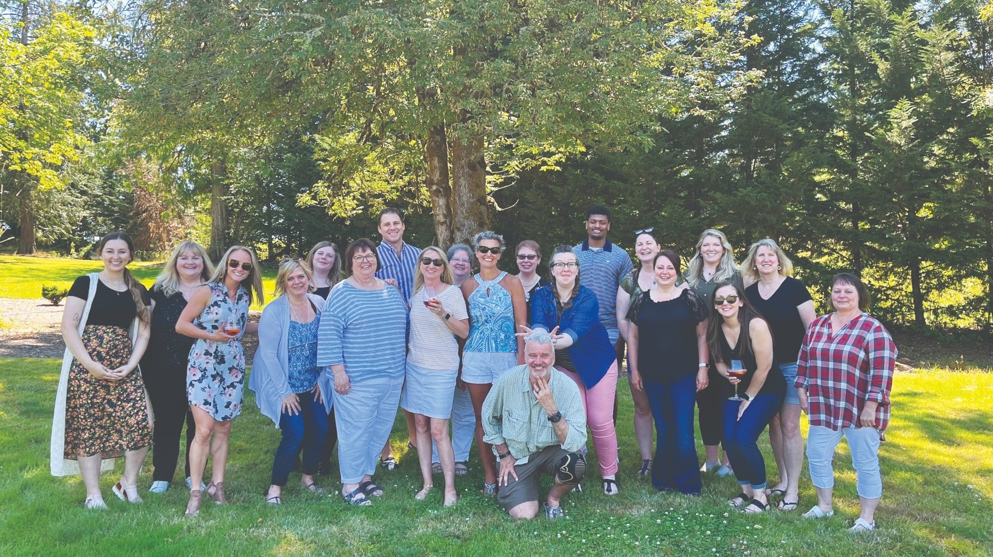 A group of adults, representing an insurance company, stands together posing for a photo in an outdoor park with trees in the background. Some are holding drinks, and all are casually dressed, reflecting the relaxed company culture.