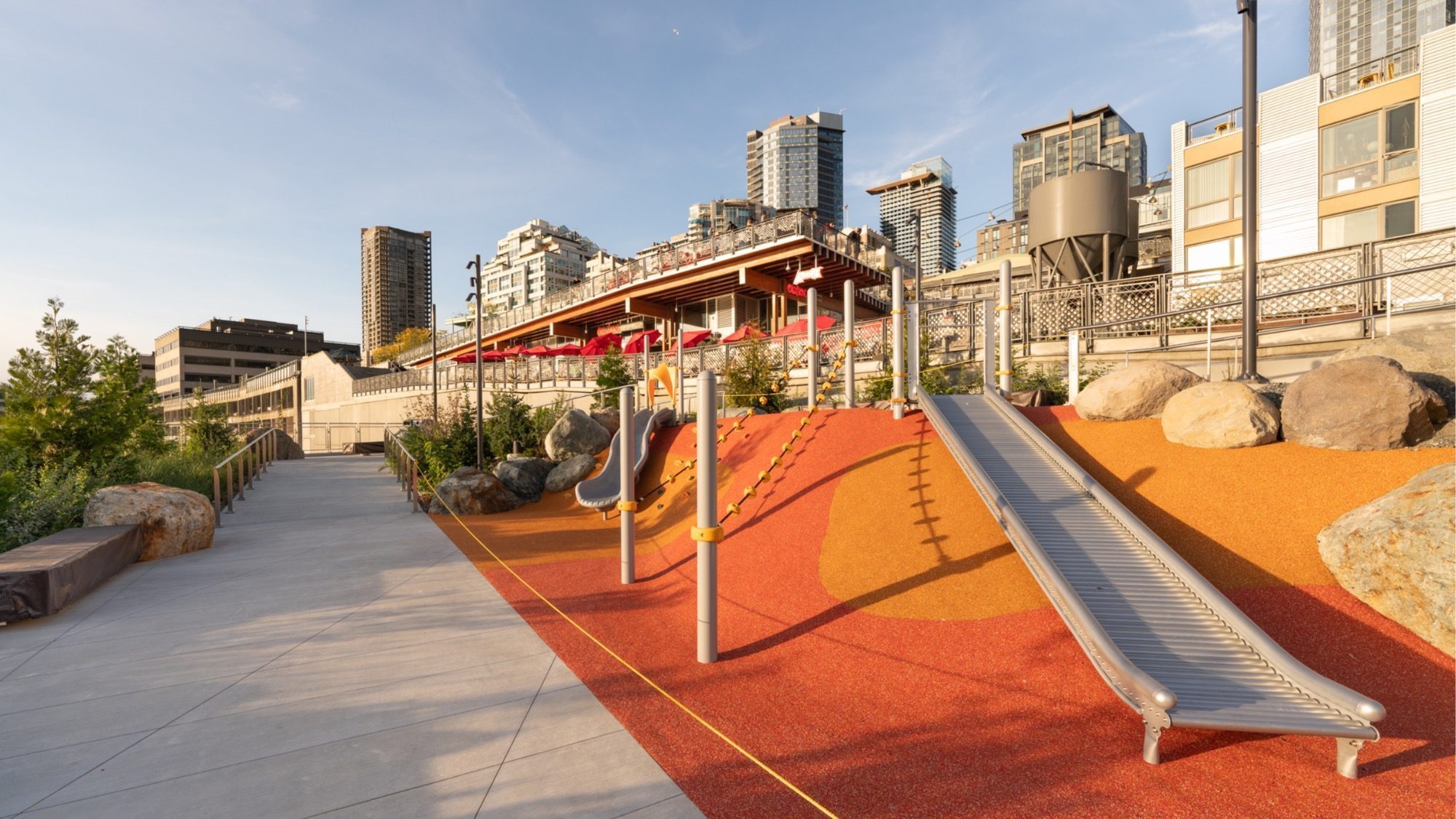 A transformative playground features slides and climbing structures on vibrant rubber flooring, nestled beside Seattle's Waterfront. Adjacent to Overlook Walk, it offers views of urban walkways and high-rise buildings in the background.