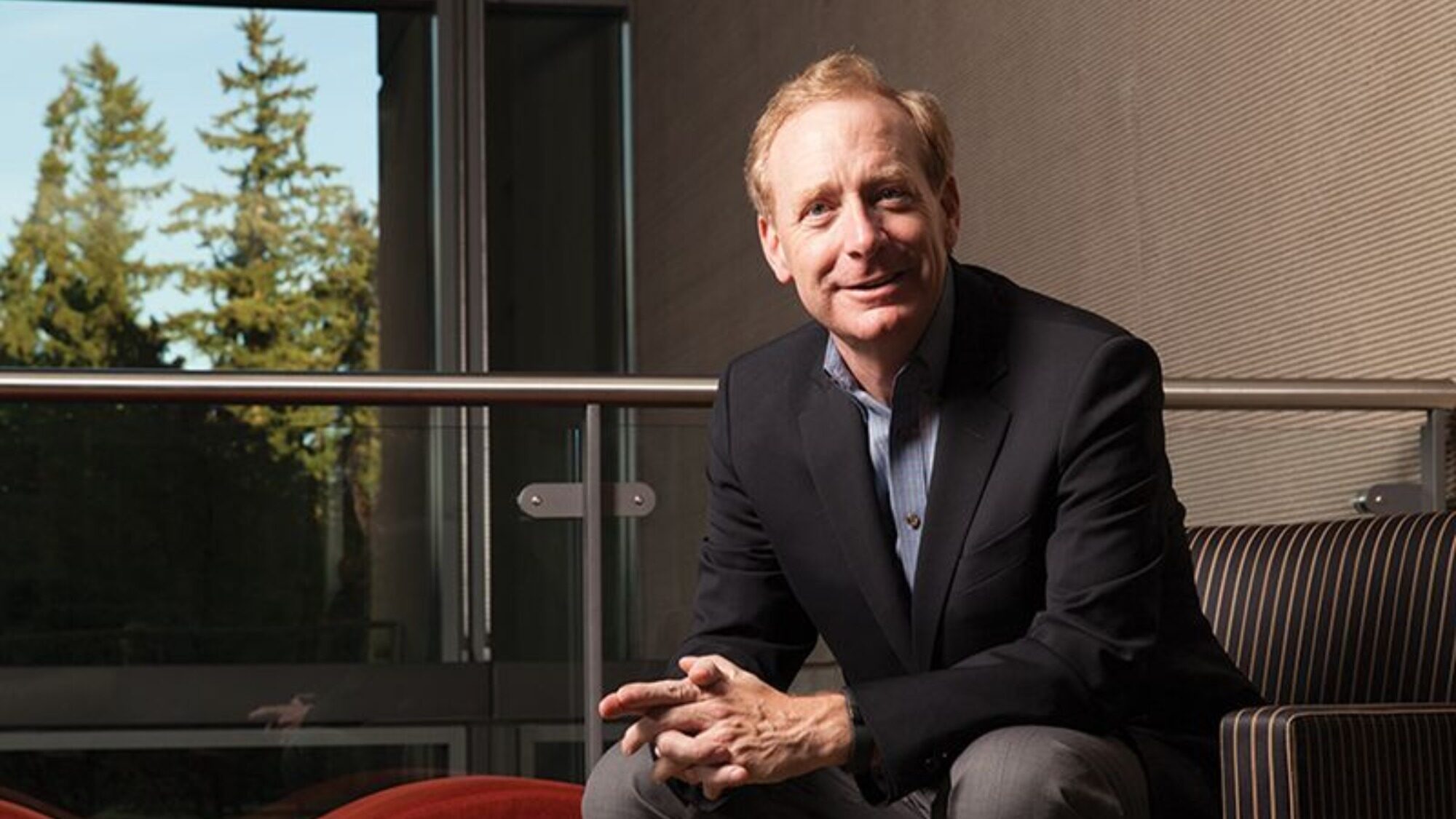 A man in a suit sits on a striped chair indoors, smiling, with a window and trees visible in the background.