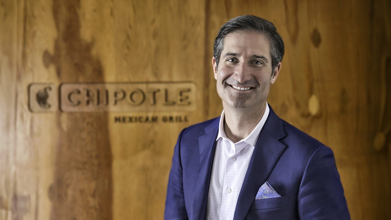 A man in a suit stands and smiles in front of a Chipotle Mexican Grill sign on a wooden wall background, exuding CEO confidence.