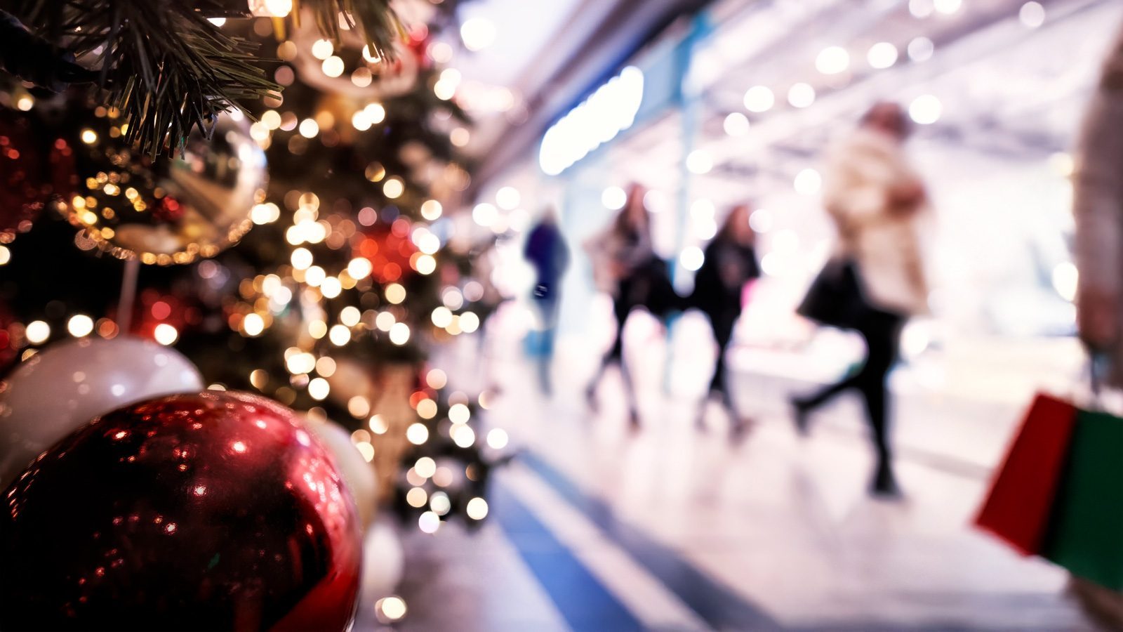 A decorated Christmas tree with red and gold ornaments graces the foreground, while blurry shoppers clutching Starbucks bags meander in the background amidst bright, festive lights.