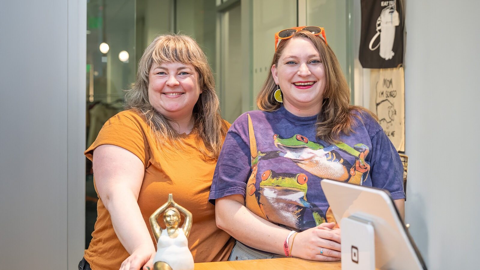 Two people smiling while sitting at a table with a small statue and a tablet. One wears an orange shirt, the other sports a colorful frog design. It's as vibrant as discovering the Curvy Cactus on a sunny stroll.