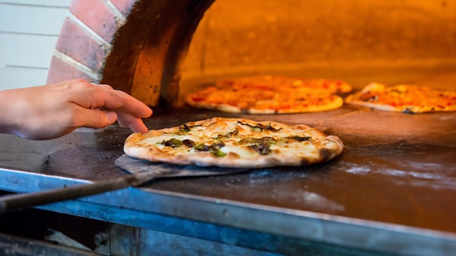 A person uses a peel to slide a freshly baked, saucy serenade of a Seattle pizza out of a brick oven, where other pizzas are also baking.