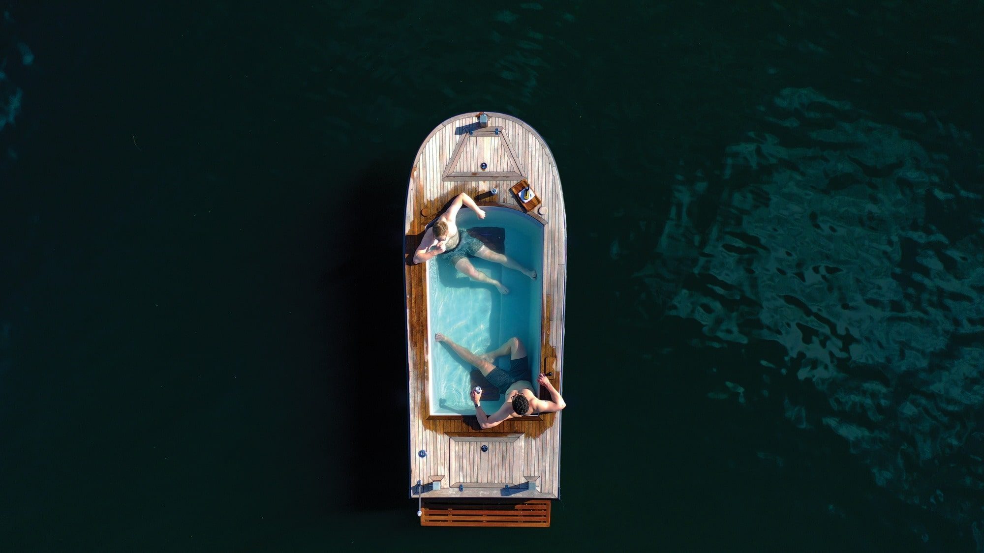Two people unwind in Seattle's hot attraction, a small wooden hot tub boat floating on dark waters. One reclines while the other sits, both savoring the soothing soak.