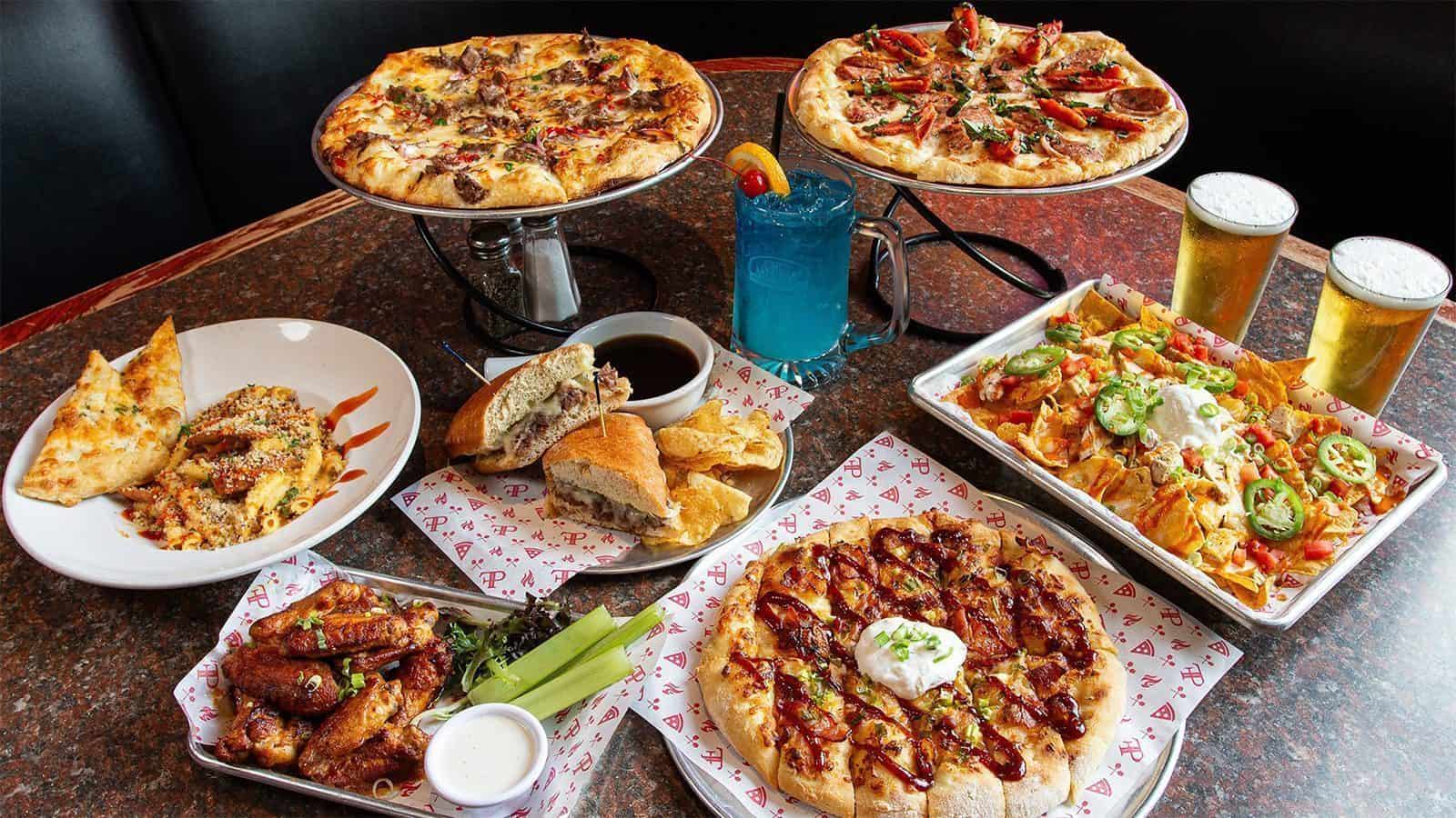 A table with various foods including two Seattle Pizzas, a salad, a bowl of pasta, wings with dipping sauce, bread, nachos, one blue drink, and two beers.