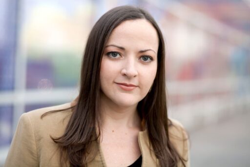 A woman with medium-length dark hair and a beige jacket looks at the camera, with a softly blurred colorful background representative of the dynamic world of finance.
