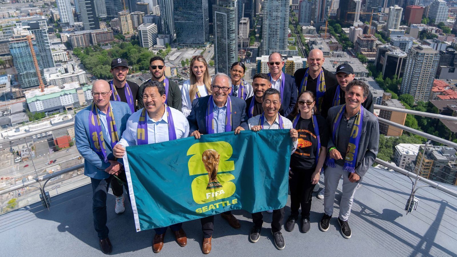 A group of people proudly holding a FIFA Seattle banner stand on a rooftop, preparing for the upcoming World Cup with the cityscape in the background.