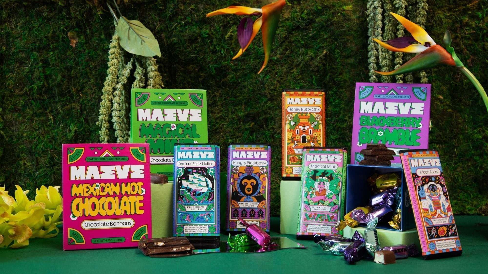 Seattle Chocolate rebrands to Maeve, offering assorted chocolate bonbon boxes with vibrant packaging on a green surface. Wrapped candies and decorative plants in the background enhance this delightful, visual treat.