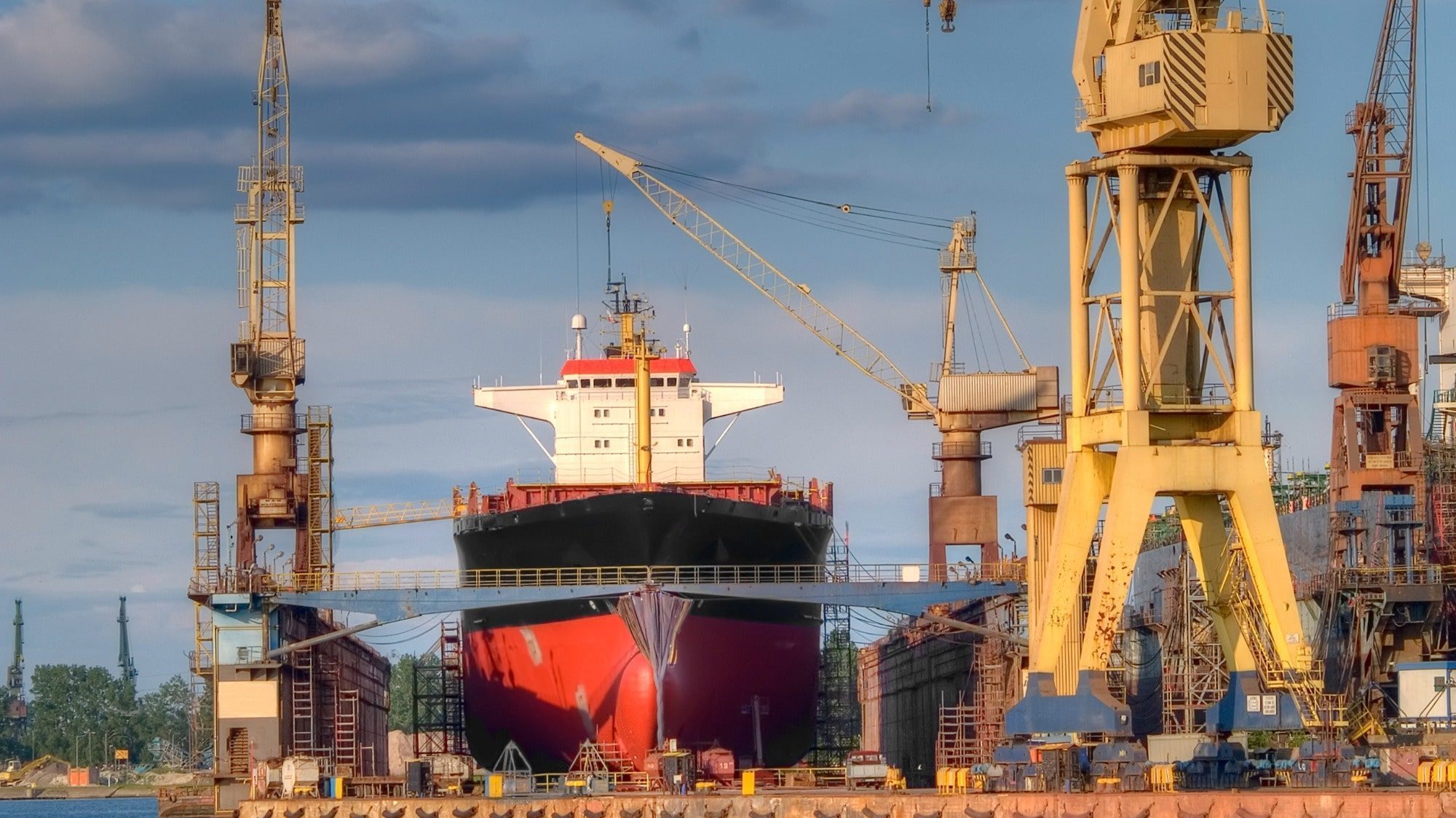 Cargo ship in dry dock at a maritime shipyard, flanked by large yellow cranes under a cloudy sky.