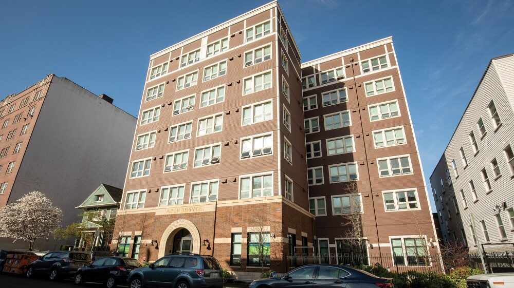 A six-story brown and beige apartment building with multiple windows, arched entryway, and parked cars lining the street in front—an example of affordable housing in Seattle amid the city's tech companies investing in Seattle.