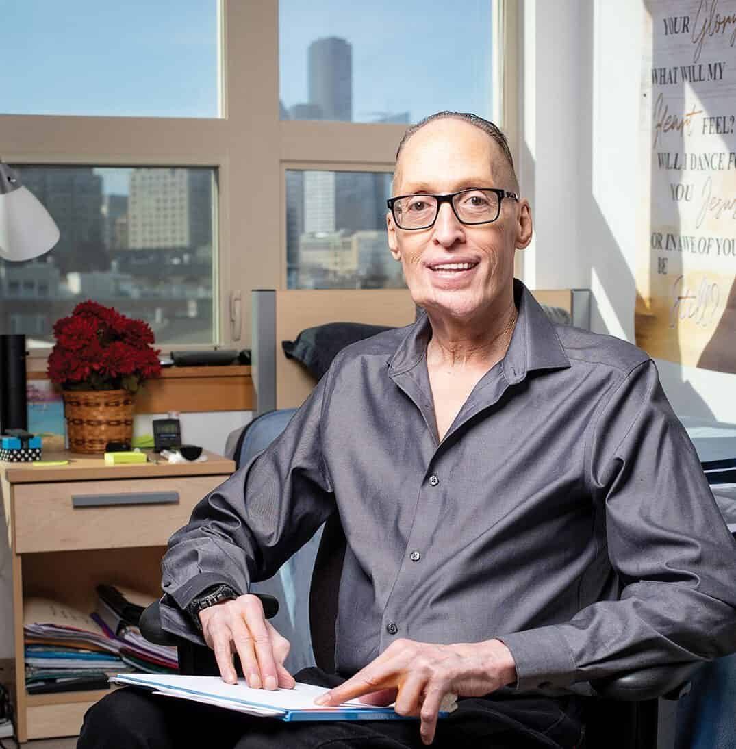 A man with glasses and a gray shirt sits in an office chair, holding an open notebook, with city buildings—home to tech companies investing in Seattle—visible through the window behind him.