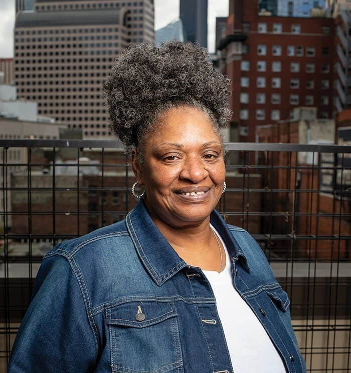 A woman with curly gray hair, wearing a denim jacket over a white shirt, smiles while standing outdoors with city buildings—home to tech companies investing in Seattle—in the background.