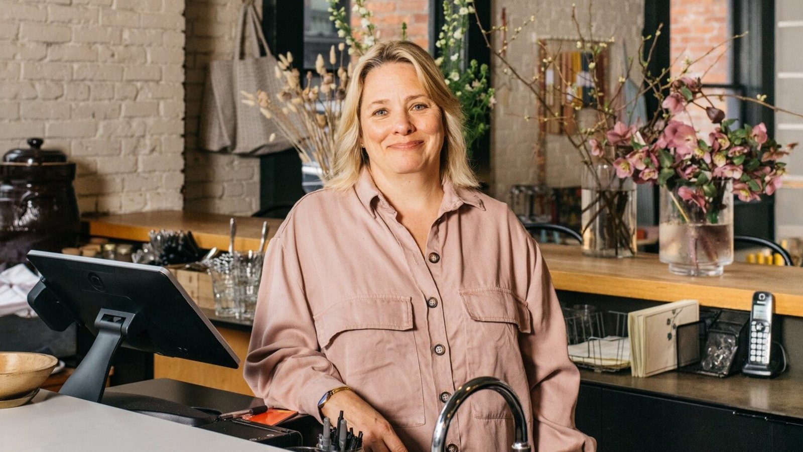 A person with blonde hair stands smiling behind a counter in a café, embodying the pioneer spirit. The setting includes flowers, a cash register, and various items on shelves.