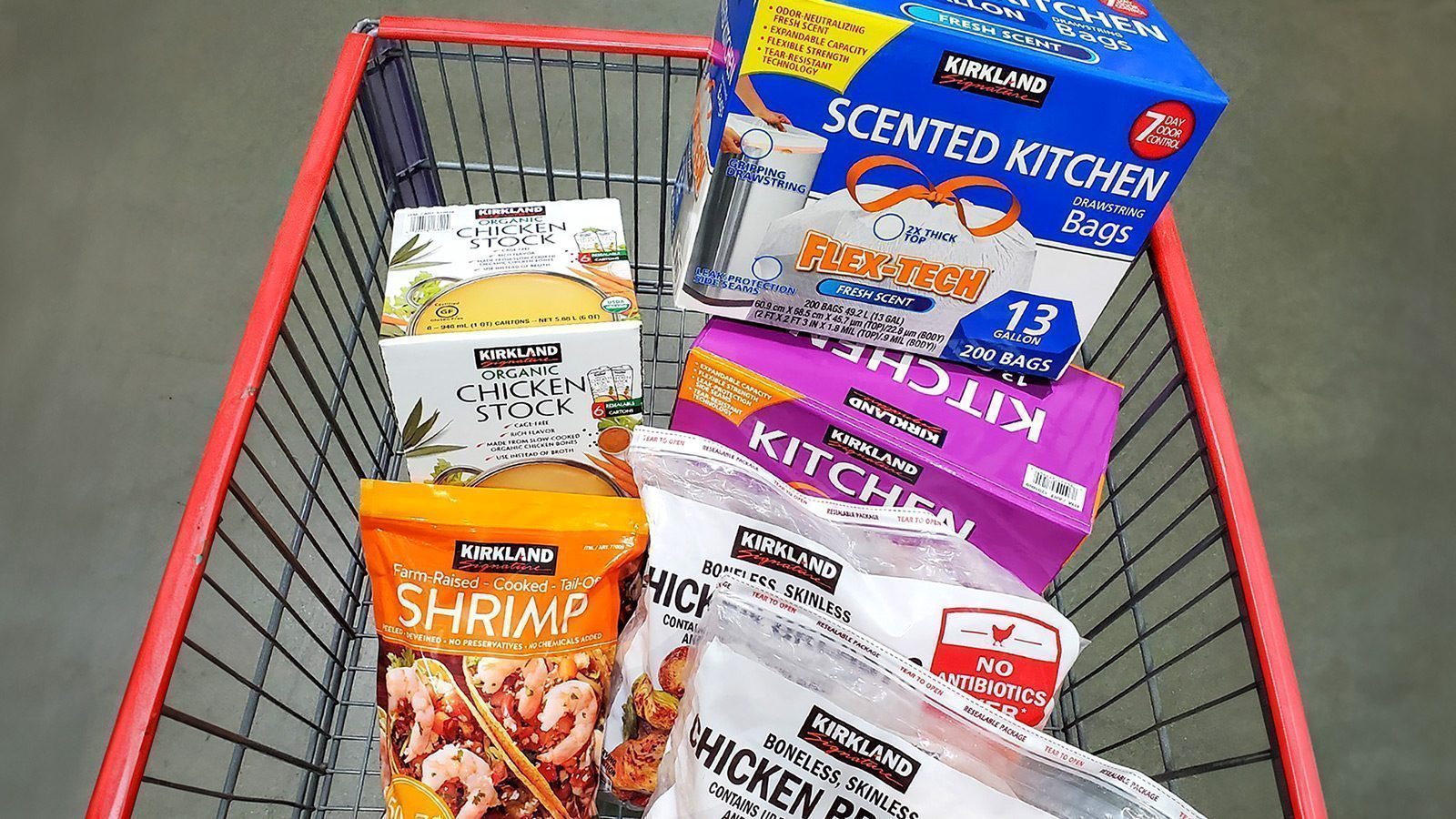 A shopping cart brimming with Kirkland Signature products delights Costco shoppers, featuring chicken stock, shrimp, chicken breasts, and kitchen bags.