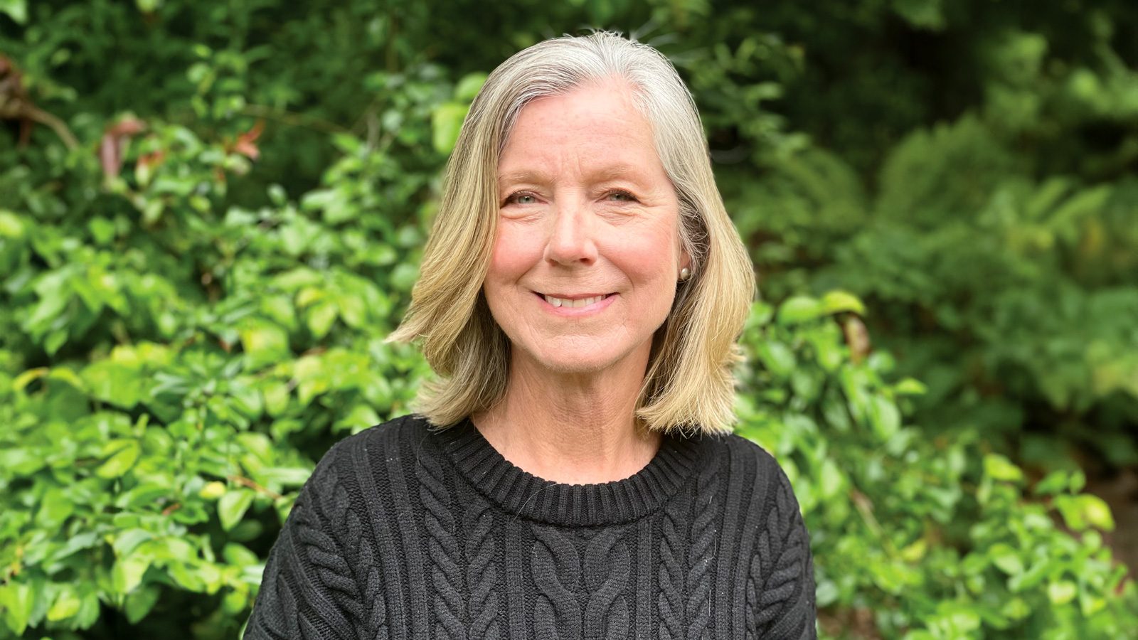 A senior captain with gray hair and a black sweater smiles warmly, surrounded by lush green foliage.