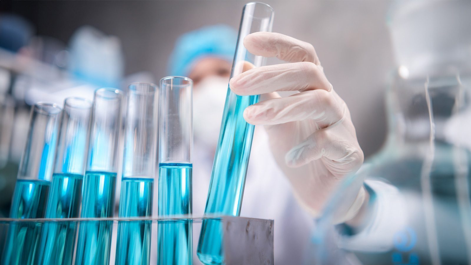 A life science scientist wearing gloves and a mask holds a test tube filled with blue liquid among several others in a laboratory setting.