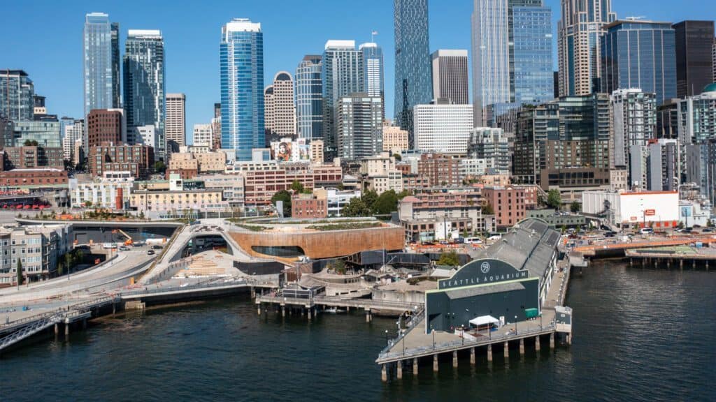 Aerial view of Seattle's waterfront showcasing the Seattle Aquarium and its New Ocean Pavilion in the foreground, with tall buildings of the downtown skyline in the background.
