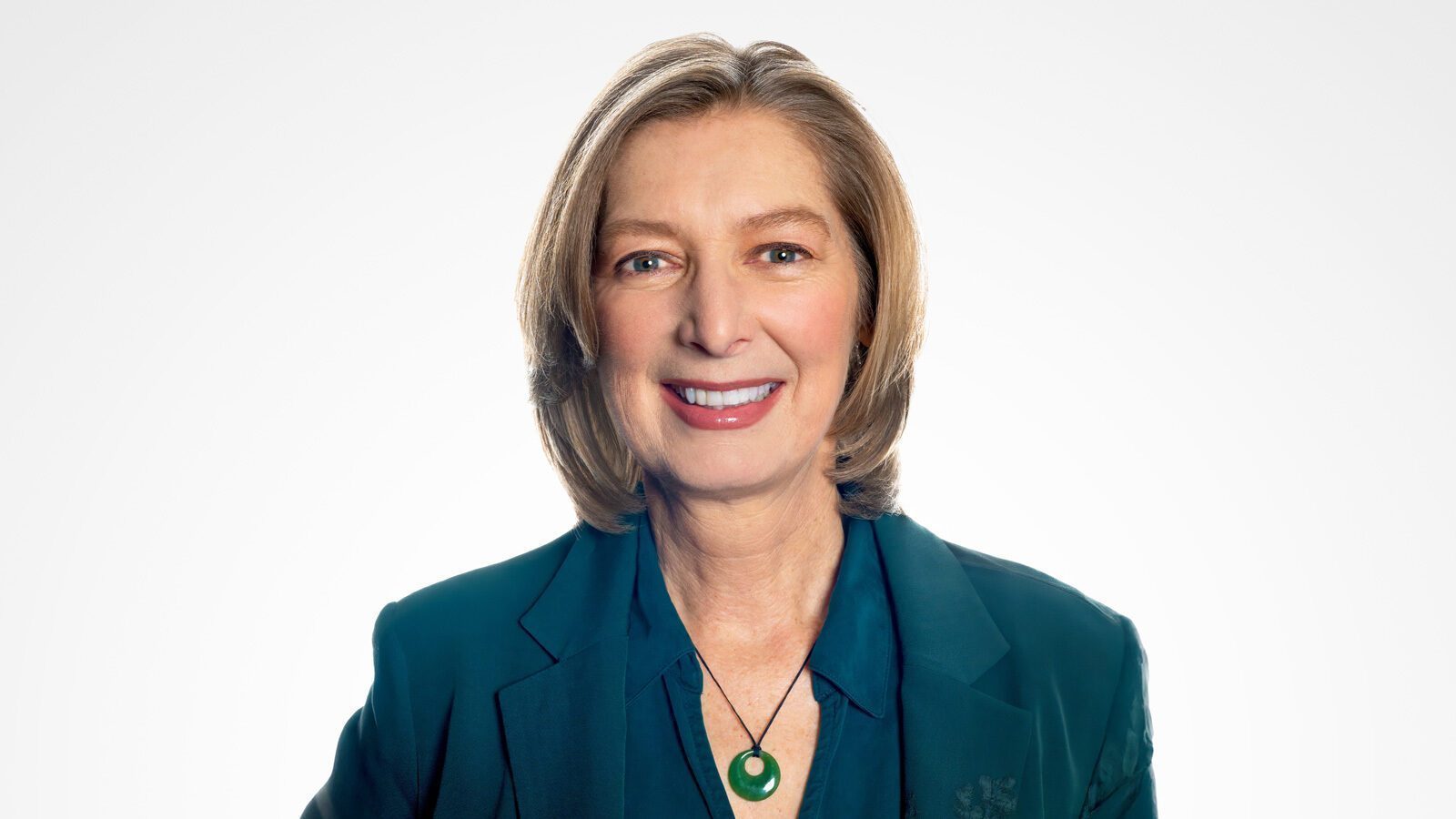 A smiling person with shoulder-length light brown hair, the new CEO, is wearing a dark teal blazer and blouse paired with a green pendant necklace. They're poised against a light background, reflecting their vibrant leadership style akin to the dynamic energy of the Seattle Aquarium.