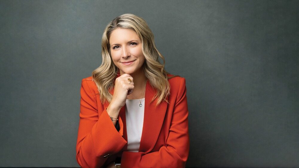 A Seed Strategist with wavy blonde hair wearing an orange blazer and white top sits at a table, smiling slightly with her hand resting under her chin against a plain gray background.