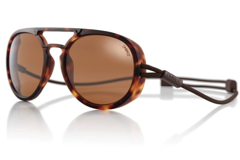 Tortoiseshell aviator sunglasses with brown tinted lenses and an adjustable strap rest on a white surface, reflecting light. The warm tones of the glasses echo the hues of a sunlit desert landscape, reminiscent of a camel's journey under the sun.