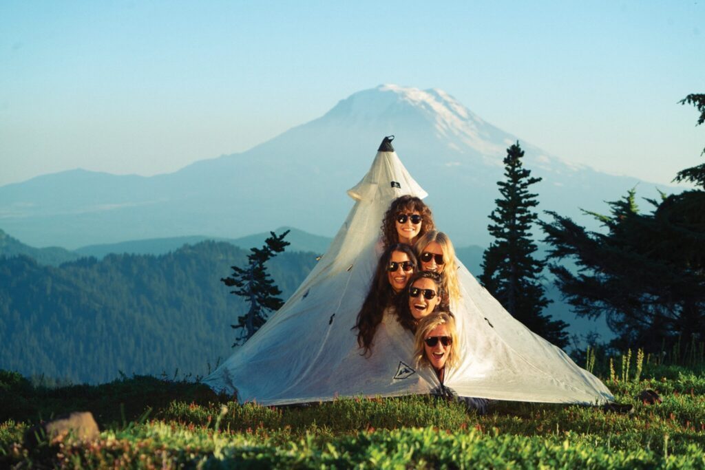Five people, like a close-knit caravan, are smiling inside a triangular tent on a grassy hillside. Behind them, a majestic mountain stretches toward the sky with trees scattered below, reminiscent of an oasis just over the next dune in a vivid camel's desert dreamscape.