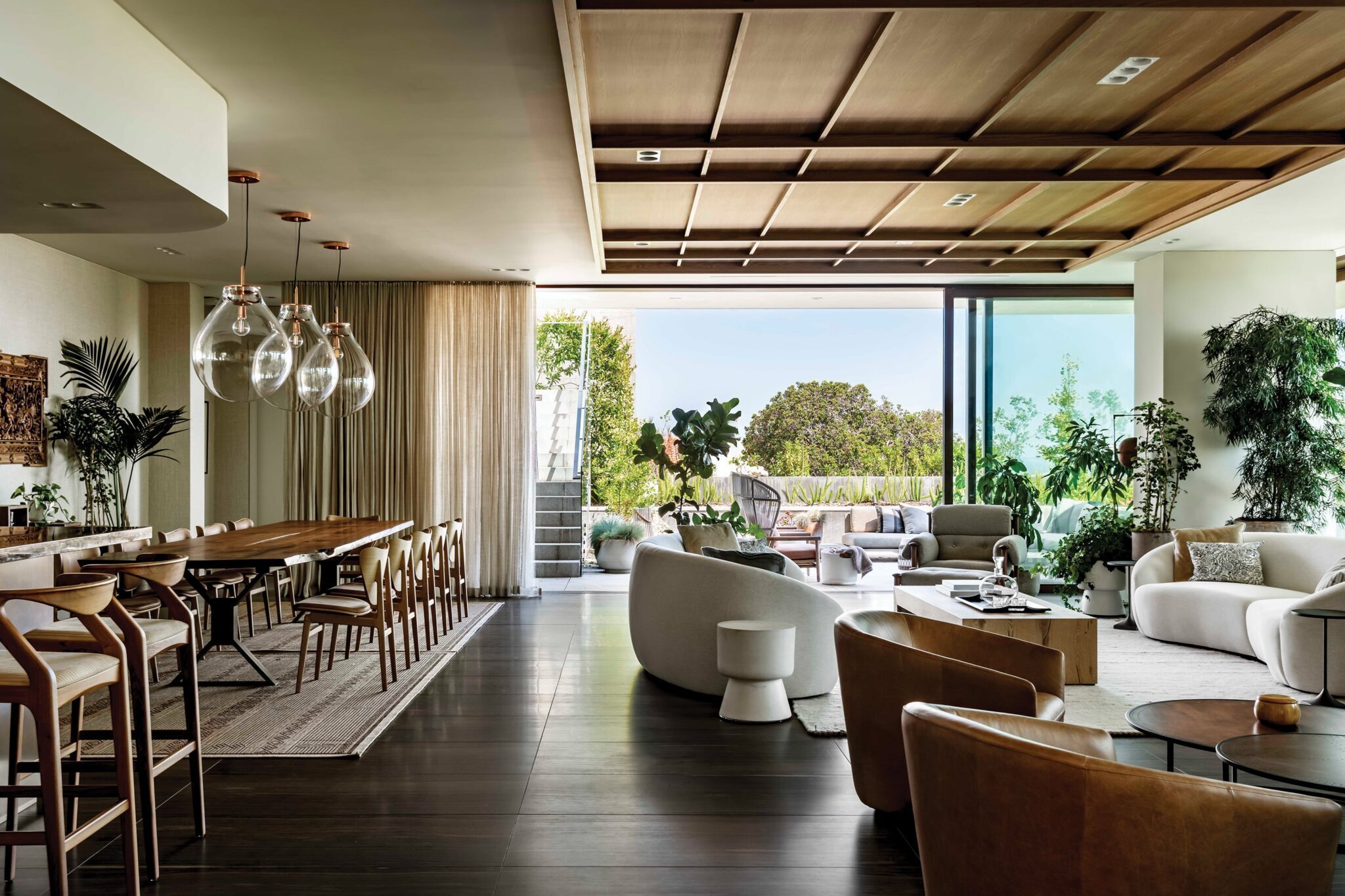 Spacious living and dining area designed by architect Kyle Gaffney, featuring modern furniture, large glass doors, and abundant greenery indoors and outdoors.