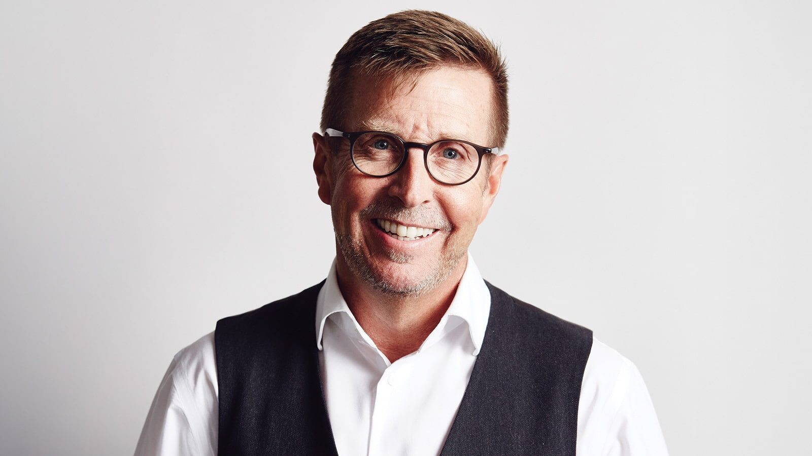 Architect Kyle Gaffney, known for his innovative designs, stands confidently in a white shirt and dark vest, his glasses adding a touch of sophistication to his smile against the plain background.