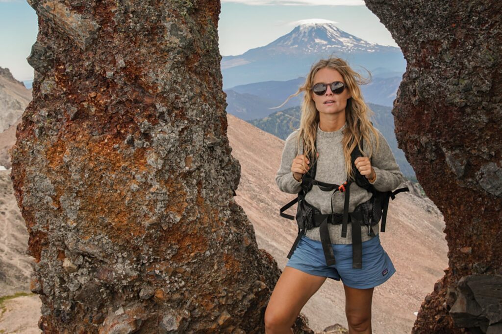 A person in hiking attire stands between rocky formations, reminiscent of desert cliffs, with a snow-capped mountain in the background.