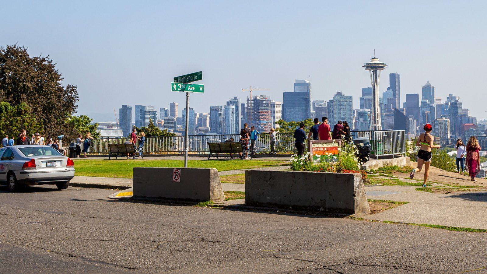 Scenic view of the Seattle skyline from a park, with people walking and signs marking 3rd Ave W. The Space Needle is visible in the background. It's a picturesque reminder that it's always the best time to hit the gym and enjoy such beautiful surroundings.