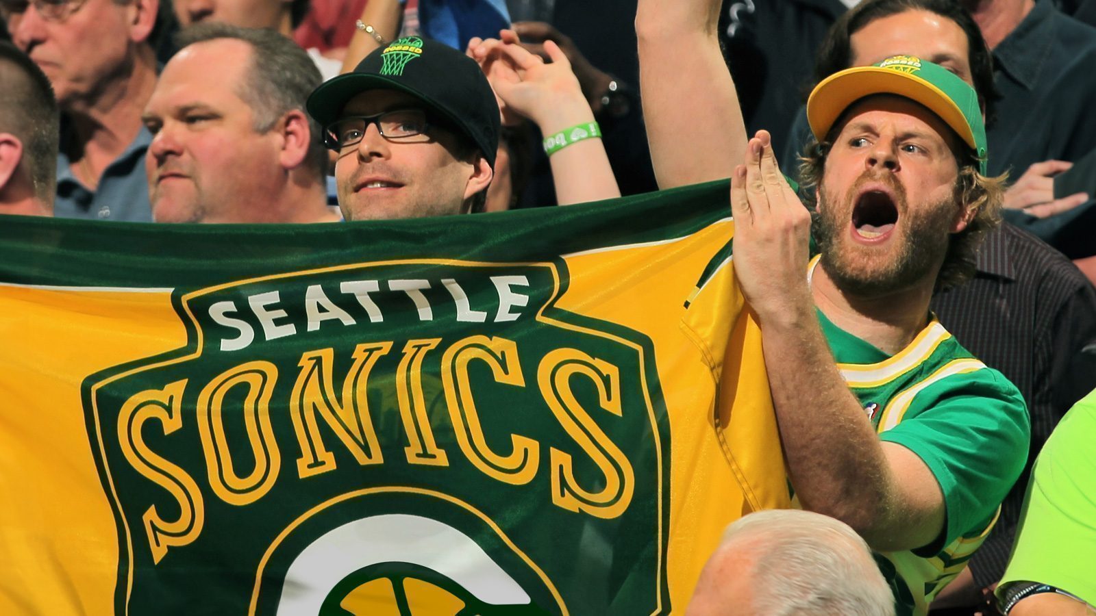 Two fans holding a "Seattle Sonics" banner at a crowded sports event. One fan is cheering loudly and the other is smiling proudly, both wearing team apparel. In the background, more Sonics supporters can be seen, adding to the vibrant atmosphere.