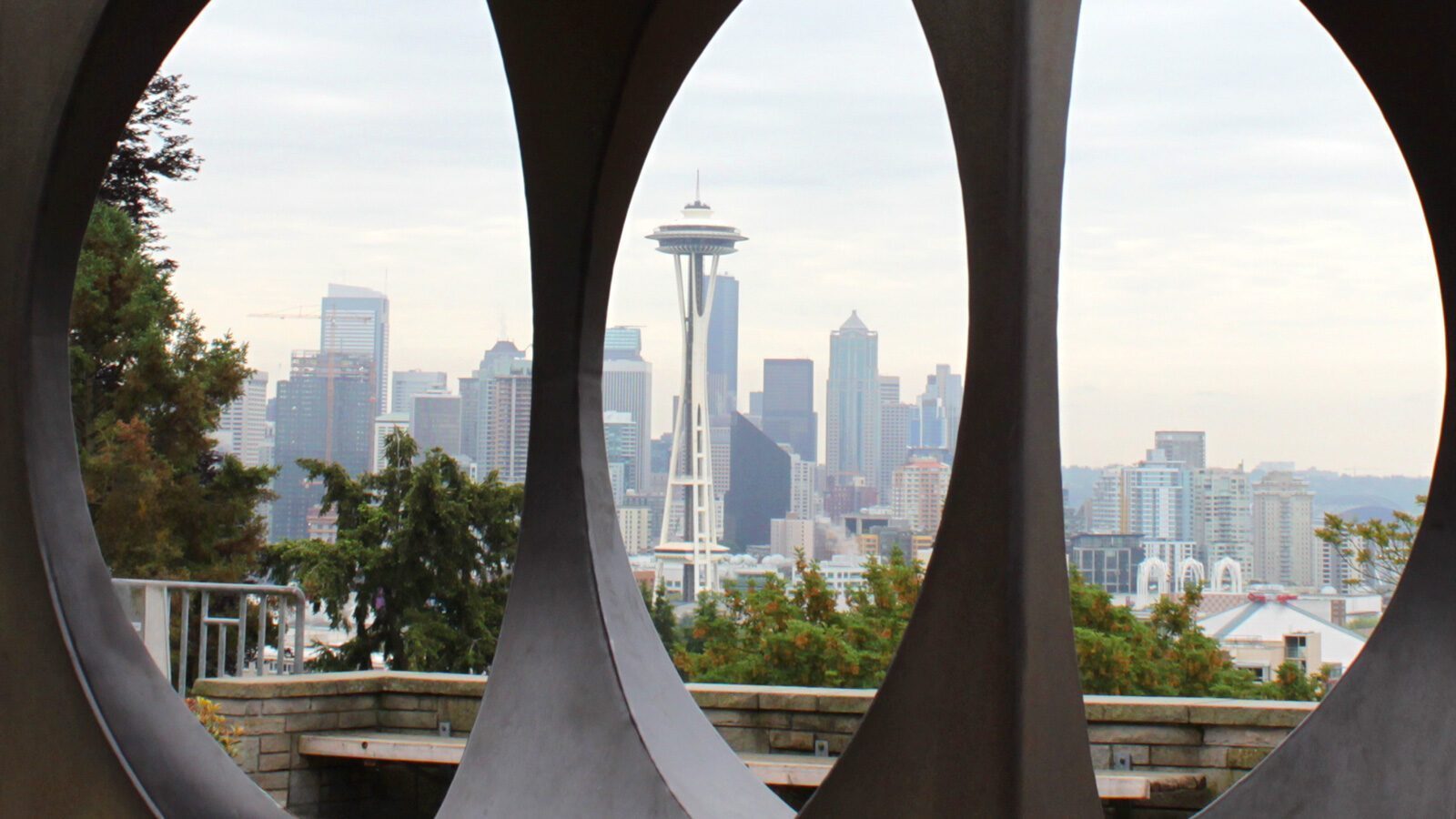 Seattle skyline featuring the Space Needle, viewed through circular cutouts in a sculpture at Seattle Center, with trees in the foreground.