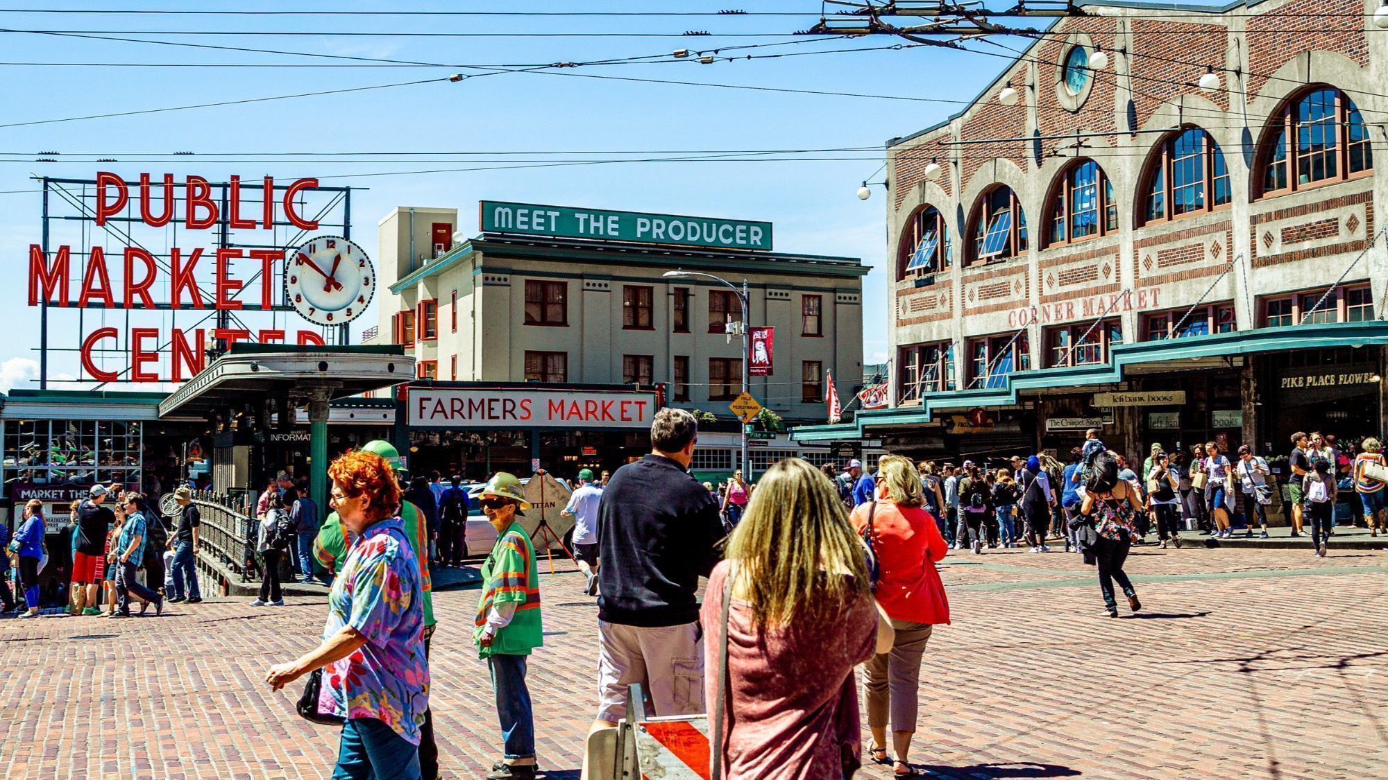 A bustling crowd walks near Pike Place Market in downtown Seattle, with visible signs for "Public Market Center" and "Farmers Market" in the background.