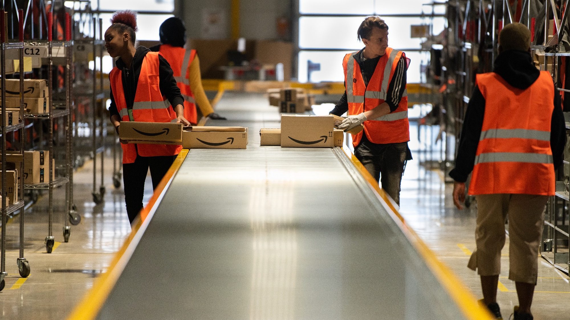 Seasonal workers in orange vests efficiently organize packages on the conveyor belt in Amazon's bustling warehouse.