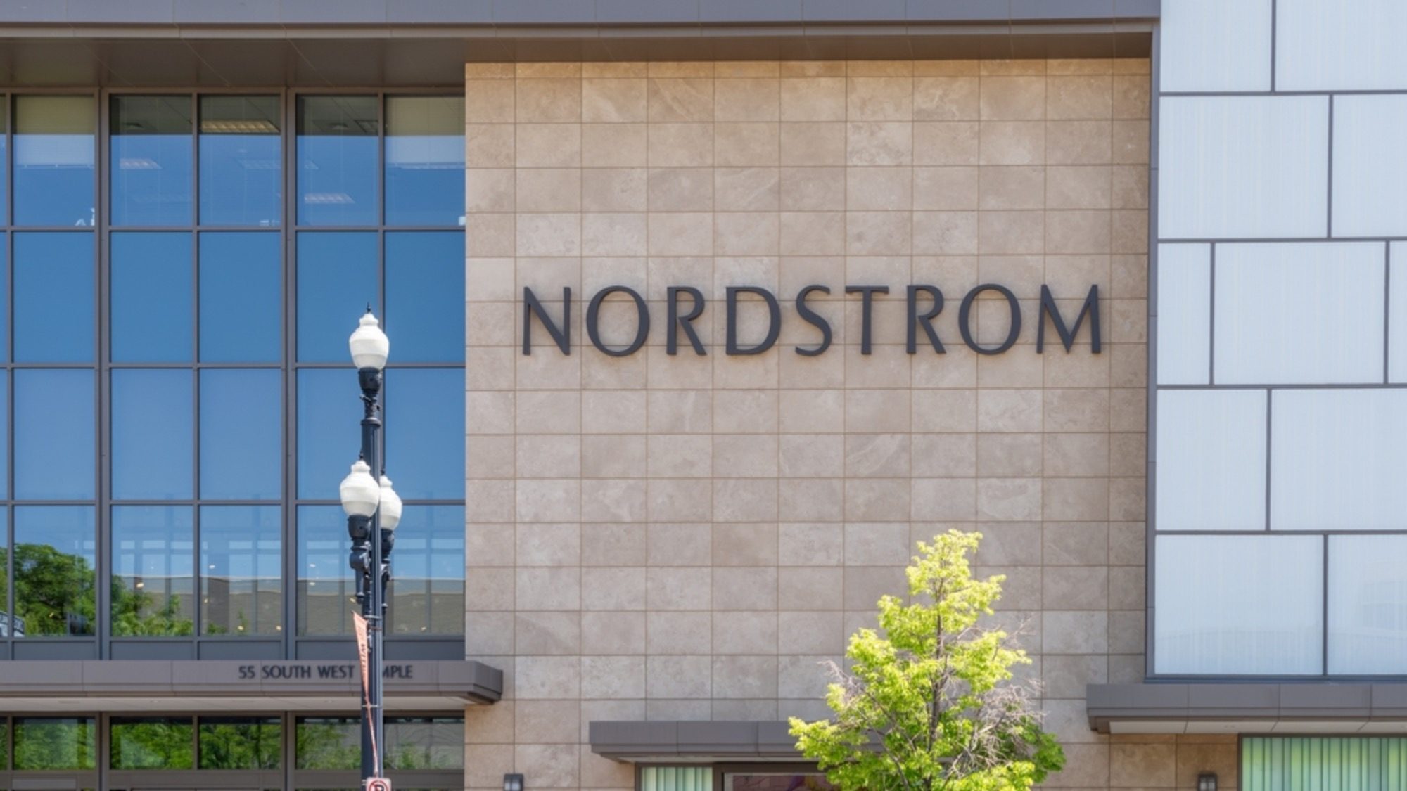 The exterior of a Nordstrom store with its name displayed on a tan-colored facade. A street lamp and a tree are visible in front of the building, perhaps witnessing conversations about ongoing merger proposals.