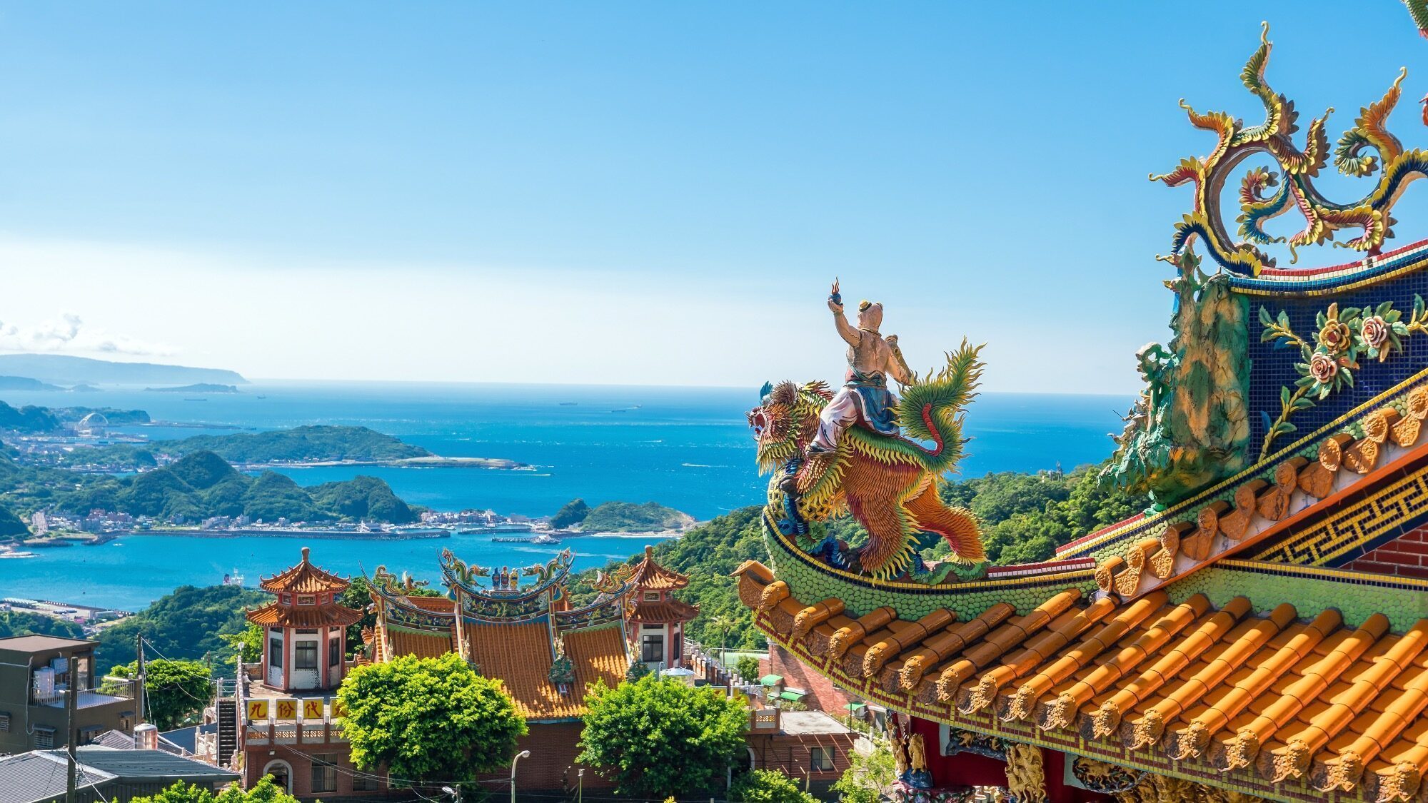 View of an ornate temple with colorful dragon sculptures on the roof overlooking a coastal town and blue ocean under a clear sky.