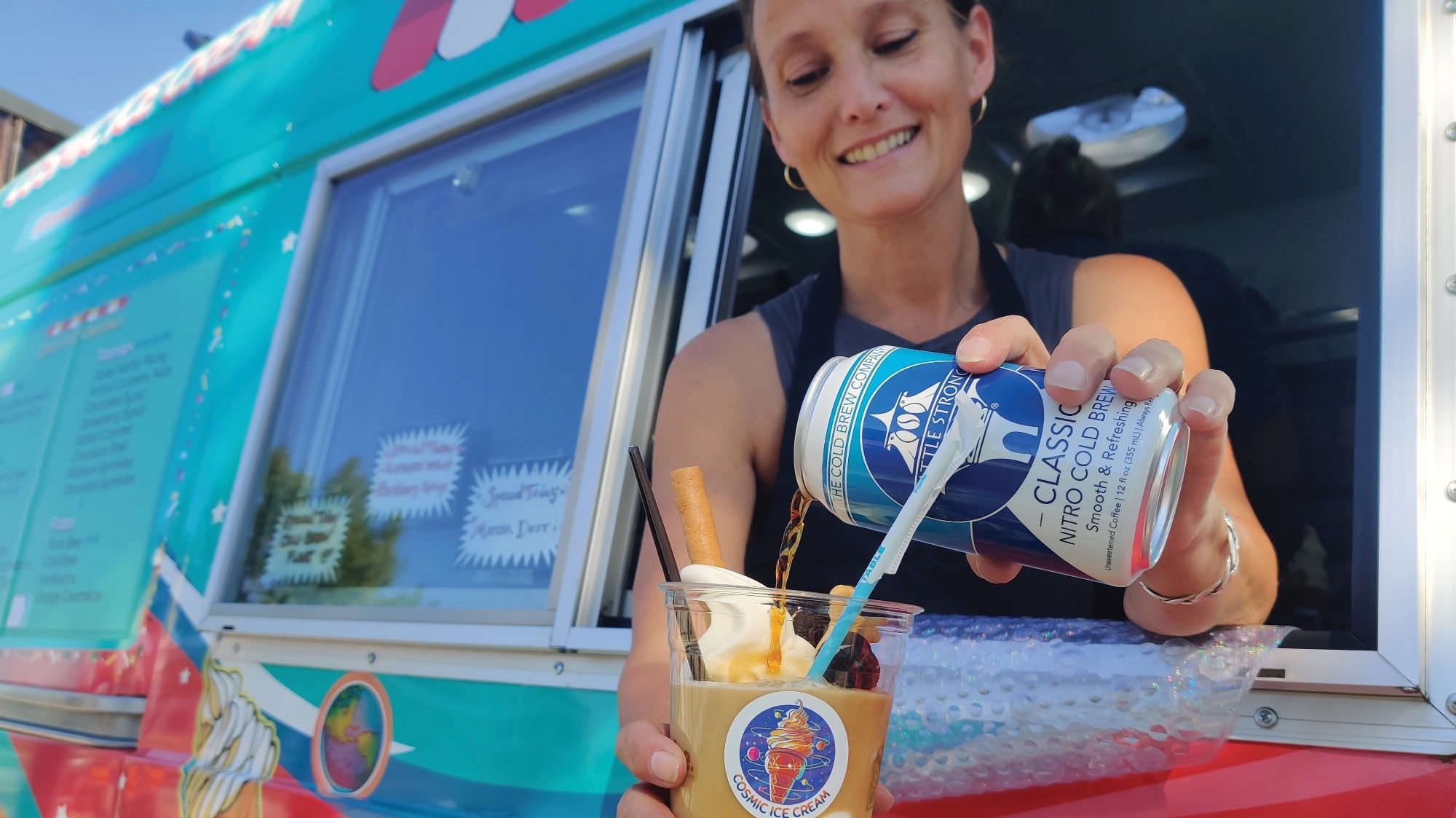 A woman pours a Seattle Strong nitro cold brew from a can into a cup, topped with whipped cream, in front of a colorful food truck.