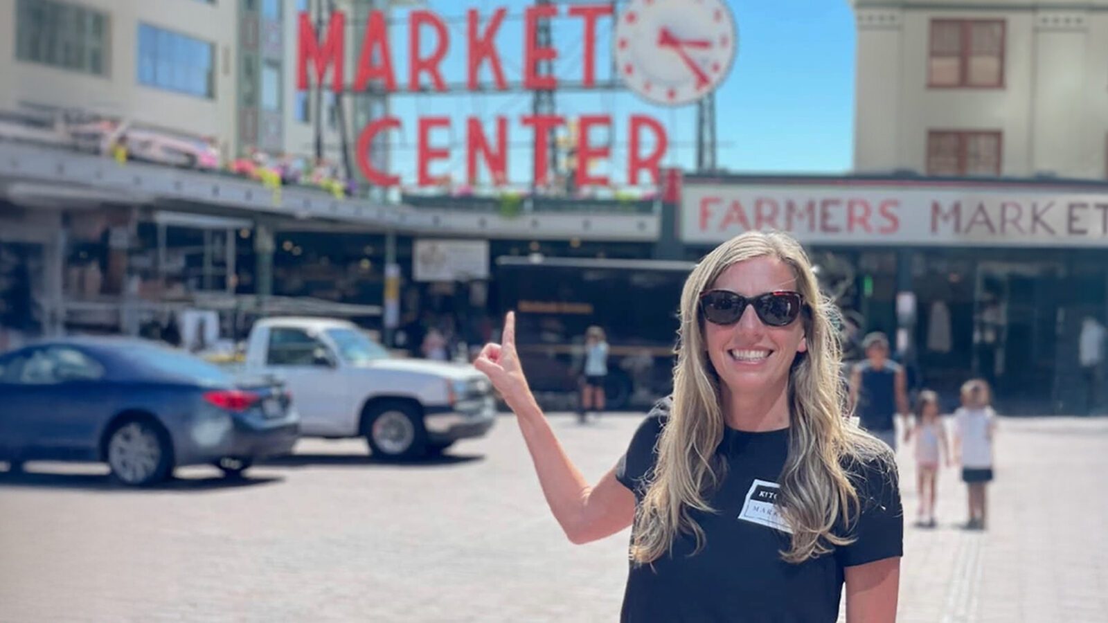 A woman wearing sunglasses and a black shirt smiles and points at the Pike Place Market Center sign in Washington state’s iconic outdoor market setting.