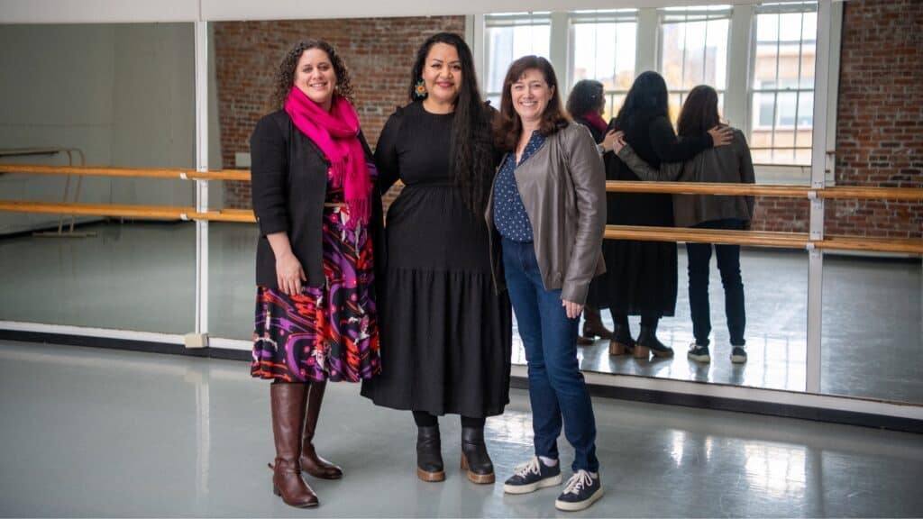Three people stand in a dance studio at Kerry Hall, surrounded by mirrors, wood floors, and large windows that flood the room with light.