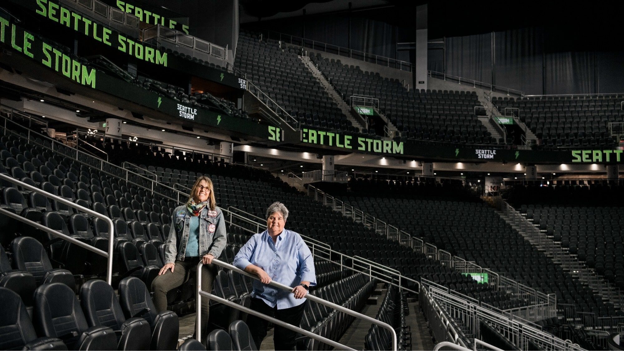 Two people stand among empty seats inside a large indoor arena with multiple digital signs displaying "SEATTLE STORM.