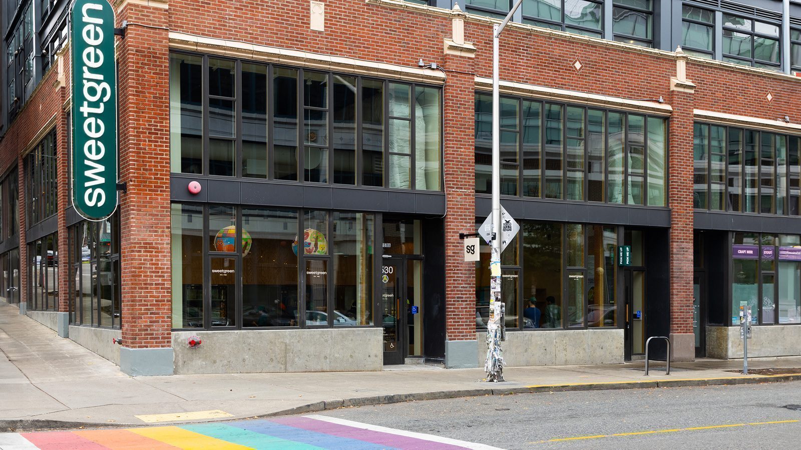 Street view of a Sweetgreen restaurant located in a corner brick building on Capitol Hill, with large windows and a rainbow crosswalk in the foreground.