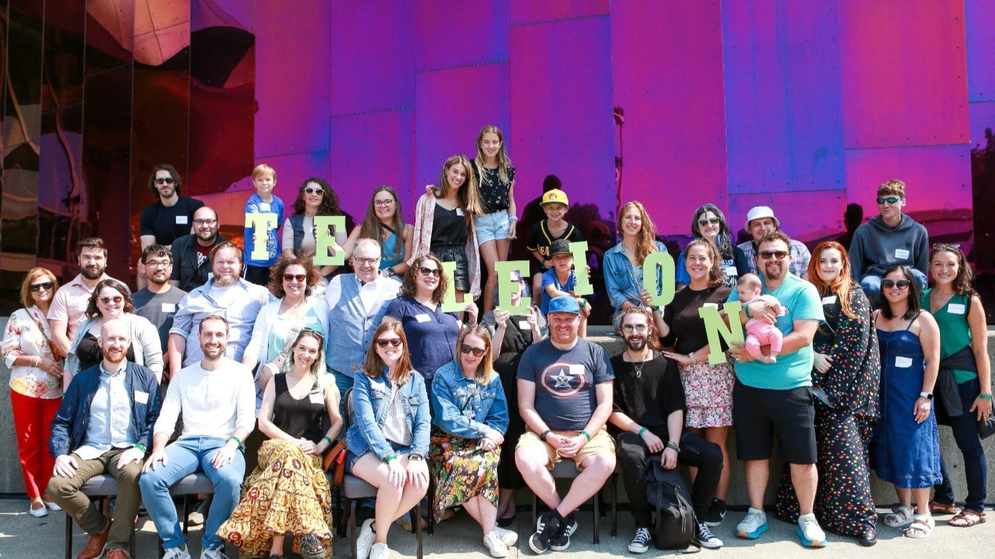 A group of people, including adults and children, pose joyfully in front of a colorful background, reminiscent of the lively spirit captured by Teleion.