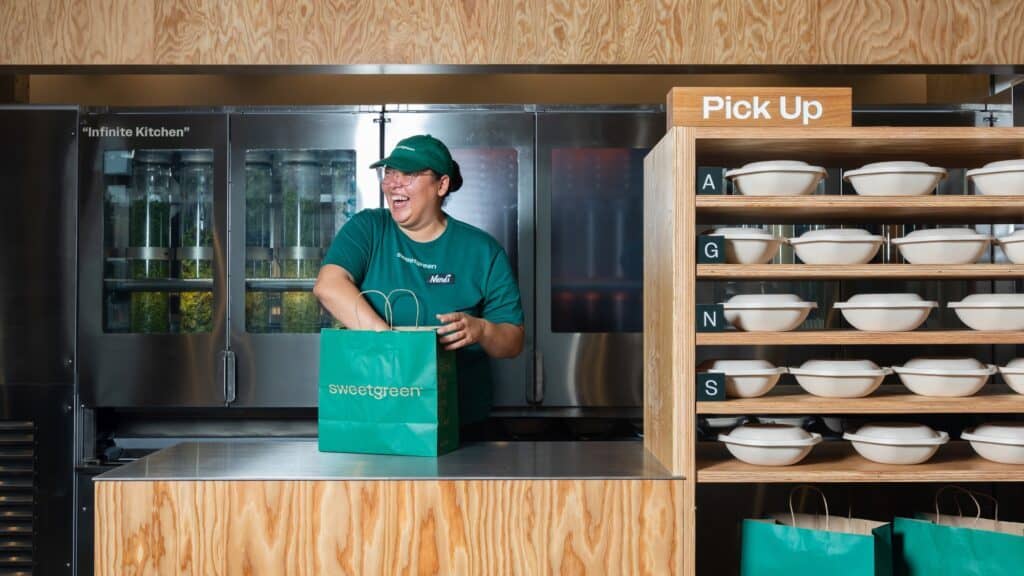 A worker in a green uniform stands behind a counter at the new Sweetgreen opening on Capitol Hill, holding a Sweetgreen bag. Shelves with packaged bowls are visible next to a "Pick Up" sign.