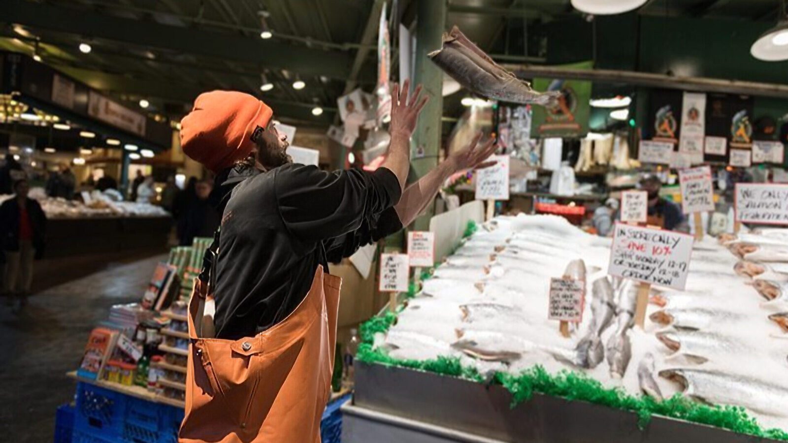 A fishmonger wearing an orange hat and apron throws a fish in the air at an indoor seafood market, celebrating the lively atmosphere as The Numbers Are In: Seattle Tourism Is Back, with various fish displayed on ice.
