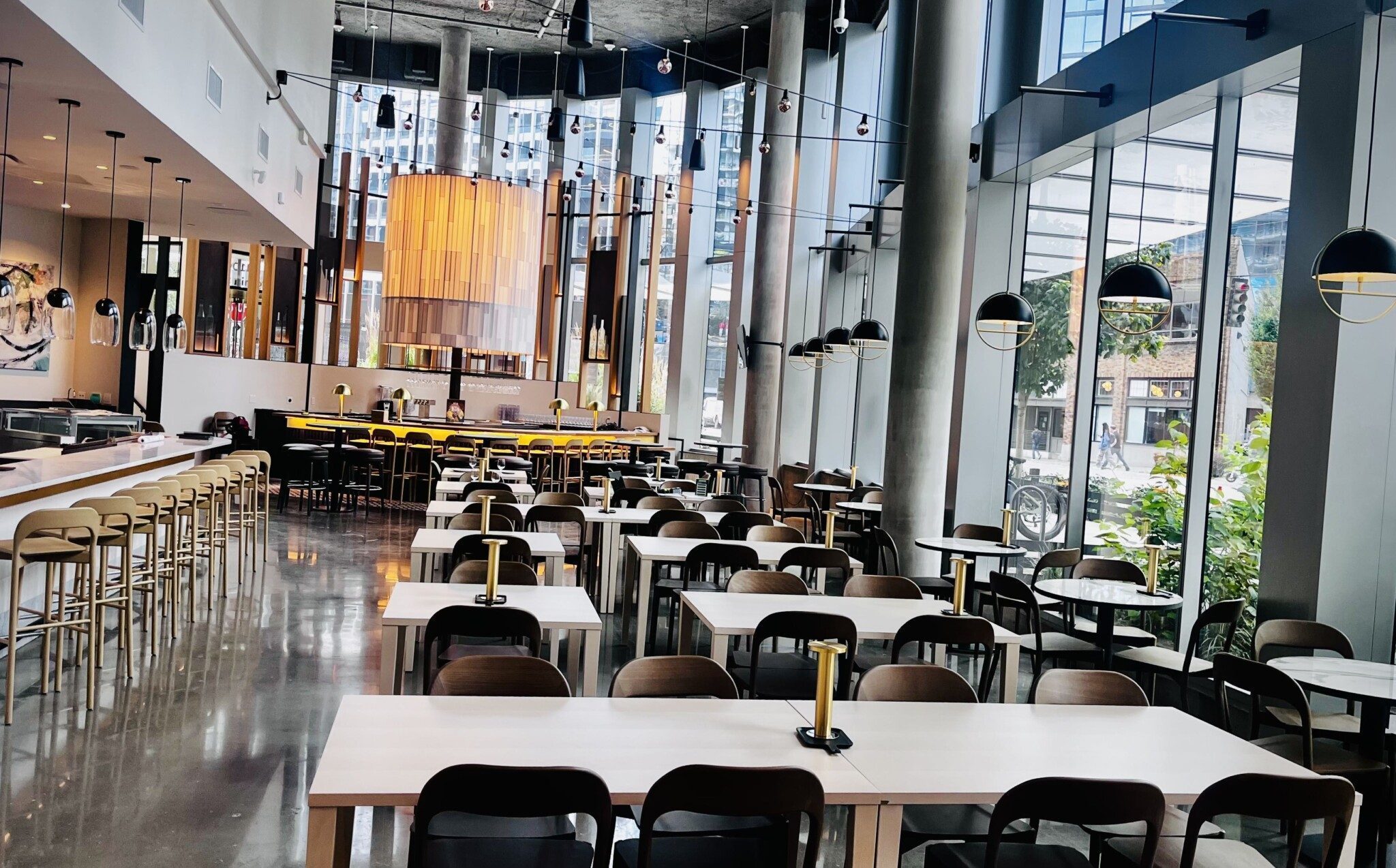 Modern, empty restaurant interior with wooden tables and chairs, pendant lighting, and large windows overlooking a street.